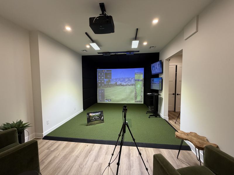 Example build style: custom golf simulator room with wood-paneled walls, tiered seating, and a tensioned impact screen showing a course view